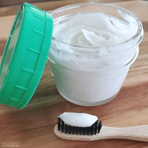 DIY coconut oil toothpaste- small jar filled with natural, homemade coconut oil toothpaste sitting on the counter and a toothbrush with toothpaste on it next to the jar